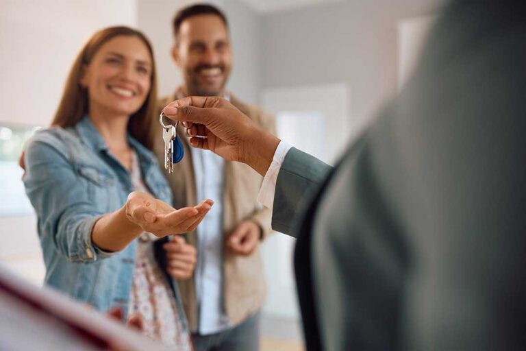 A smiling couple reviews documents with a clipboard inside a bright home filled with moving boxes.